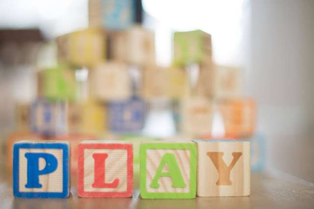 Depth-of-field photography of P-L-A-Y wooden letter. The wooden letter 'P-L-A-Y' is positioned in the foreground of the image, sharply focused and occupying the center of the frame. The background gradually blurs out, creating a shallow depth of field effect. The letter is made of wood and has a natural texture, with visible grains and a smooth finish. The lighting highlights the contours of the letter, casting subtle shadows. The overall composition conveys a playful and tactile aesthetic